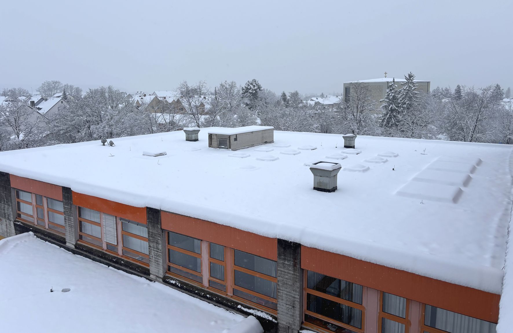 Flachdach eines Gebäudes mit mehreren Lüftungsöffnungen, komplett mit Schnee bedeckt, im Hintergrund schneebedeckte Bäume und Häuser