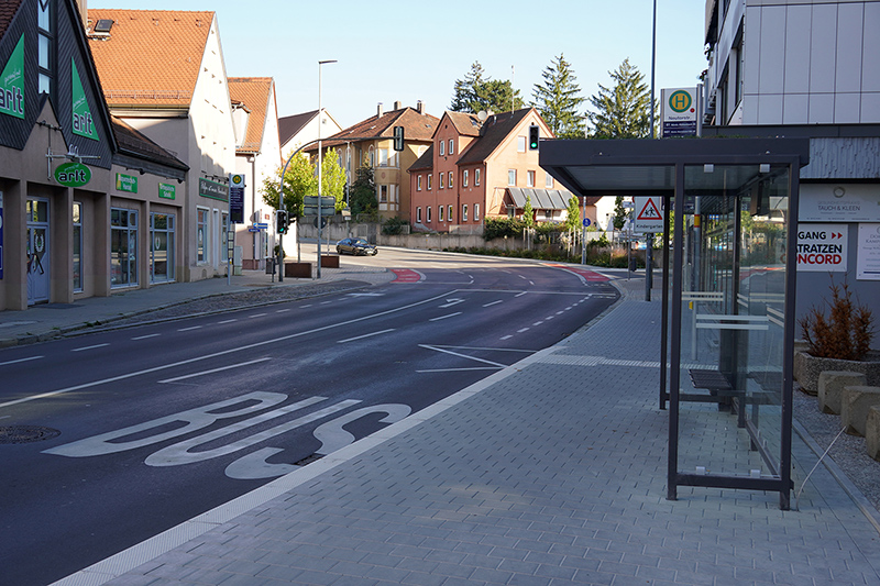 Bushaltestelle mit gläsernem Wartehäuschen am Rand einer breiten Straße. Auf dem Asphalt ist das Wort 'BUS' in großen weißen Buchstaben aufgemalt. Links stehen mehrere Geschäfte mit Schaufenstern, rechts sind Wohnhäuser und ein Schild mit Fahrplaninformationen sichtbar. Im Hintergrund verlaufen die Straße und Gehwege durch ein Wohngebiet mit Bäumen.