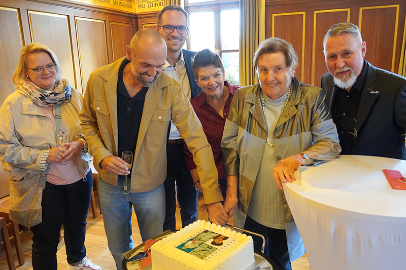 Eine Gruppe von Menschen steht um einen rechteckigen Geburtstagskuchen mit weißer Glasur. Zwei Personen schneiden gemeinsam den Kuchen an. Auf dem Kuchen ist ein Foto abgebildet. Die Menschen lächeln und halten teilweise Gläser in der Hand. Im Hintergrund sind Holzvertäfelungen und ein Fenster zu sehen.