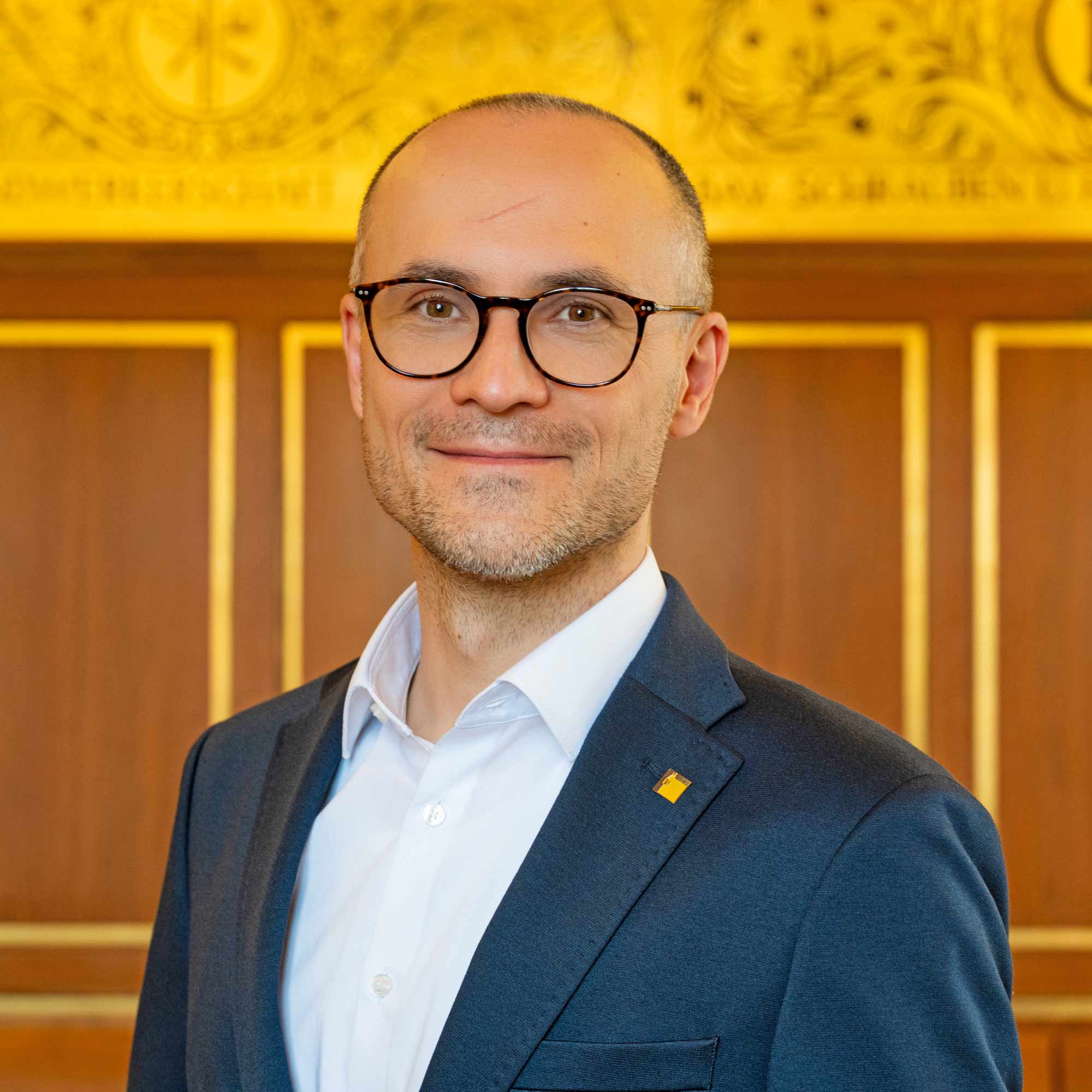 EIn Mann im blauen Anzug steht vor einer Wand mit einem Fries. Er trägt eine Brille, lächelt und hat einen Pin am Sakkokragen.