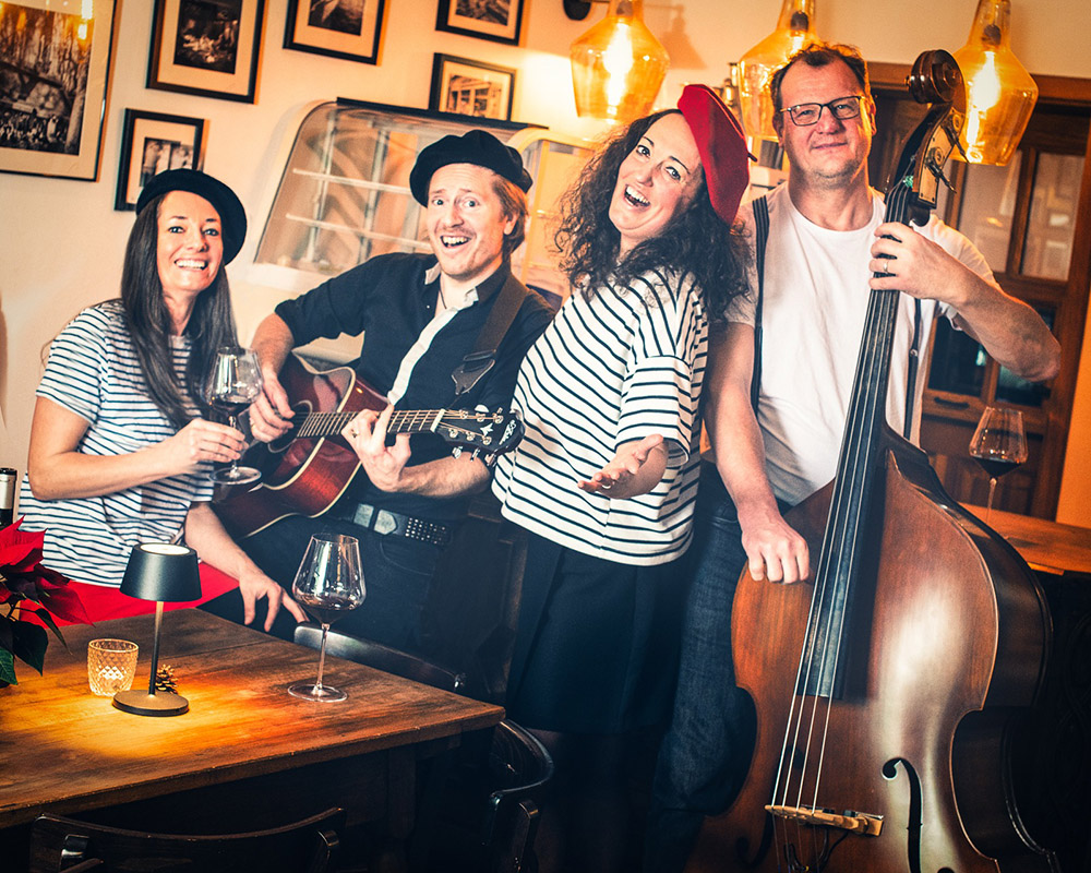 Vier Personen in französisch inspirierten Outfits mit Baskenmützen, eine spielt Gitarre, eine Kontrabass, zwei Frauen tragen schwarz-weiß gestreifte Oberteile, sie stehen in einem gemütlichen Raum mit warmem Licht