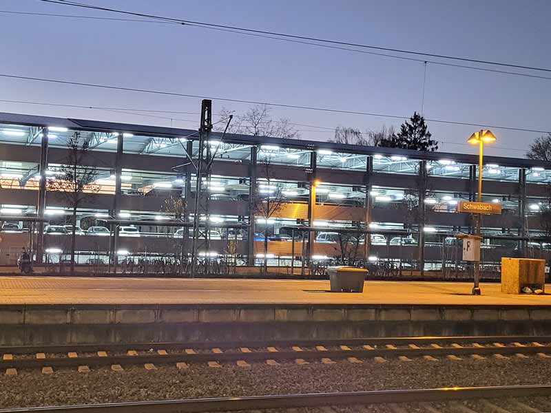 Ein Bahnsteig im Abendlicht, an dem die Beschilderung "Schwabach" zu lesen ist. Im Hintergrund sieht man ein Parkhaus, an dem die Beleuchtung eingeschaltet ist, sowie einen nächtlichen Himmel.