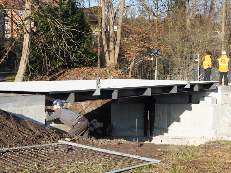 Bauarbeiter montiert eine Fahrbahnplatte auf einem Betonfundament, zwei weitere Personen mit Warnwesten stehen daneben.