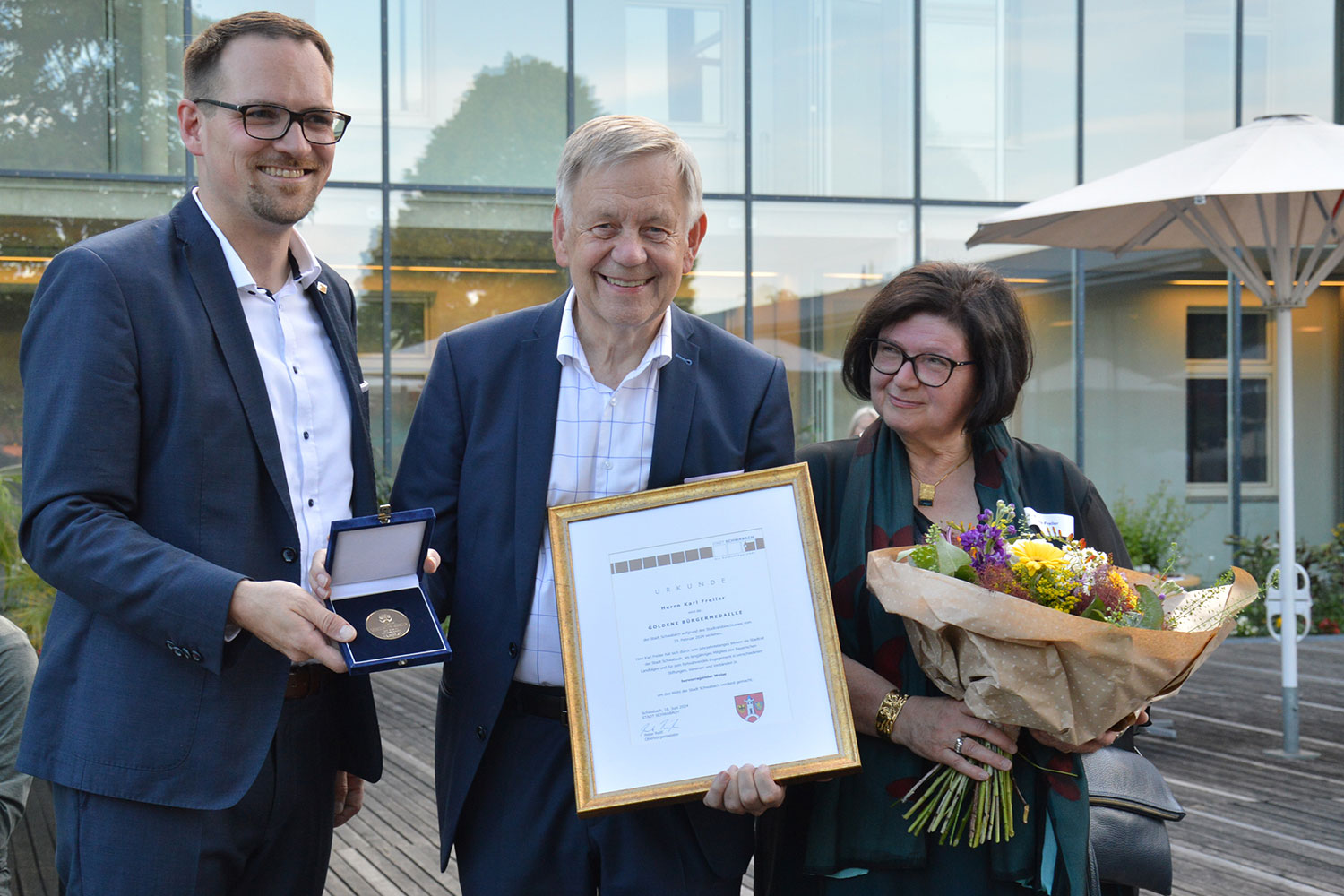Ein Herr im Anzug, Karl Freller, lächelt in die Kamera und hält eine goldene Medaille in der Hand. Links daneben Oberbürgermeister Reiß, ebenfalls im Anzug und lächelnd. Monika Freller steht rechts daneben und hält einen Blumenstrauß.