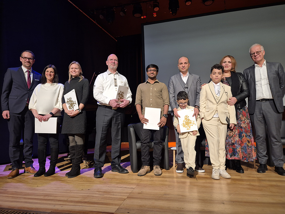 Eine Gruppe von Menschen steht nebeneinander auf einer Bühne vor einem dunklen Hintergrund. Einige halten Urkunden oder Dokumente in den Händen. Die Personen tragen unterschiedliche Kleidung, darunter Anzüge, Kleider und Pullover. Eine Frau trägt ein langes, bunt gemustertes Kleid. Die Gruppe besteht aus Erwachsenen und Kindern, die in einer Reihe stehen oder sitzen.