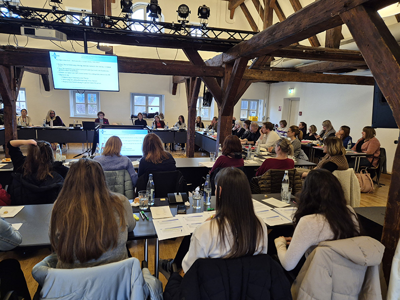 In einem großen, hellen Raum mit sichtbaren Holzbalken an der Decke sitzen zahlreiche Personen an mehreren Tischen, die zu einem großen Rechteck zusammengestellt sind. Die Personen sind überwiegend von hinten zu sehen und konzentrieren sich auf zwei große Bildschirme, die in der Mitte des Raums aufgestellt sind. Auf den Tischen liegen Papiere, Notizblöcke und Wasserflaschen. Die Fenster im Raum lassen Tageslicht herein, und die Atmosphäre wirkt wie bei einer Konferenz oder einem Meeting.