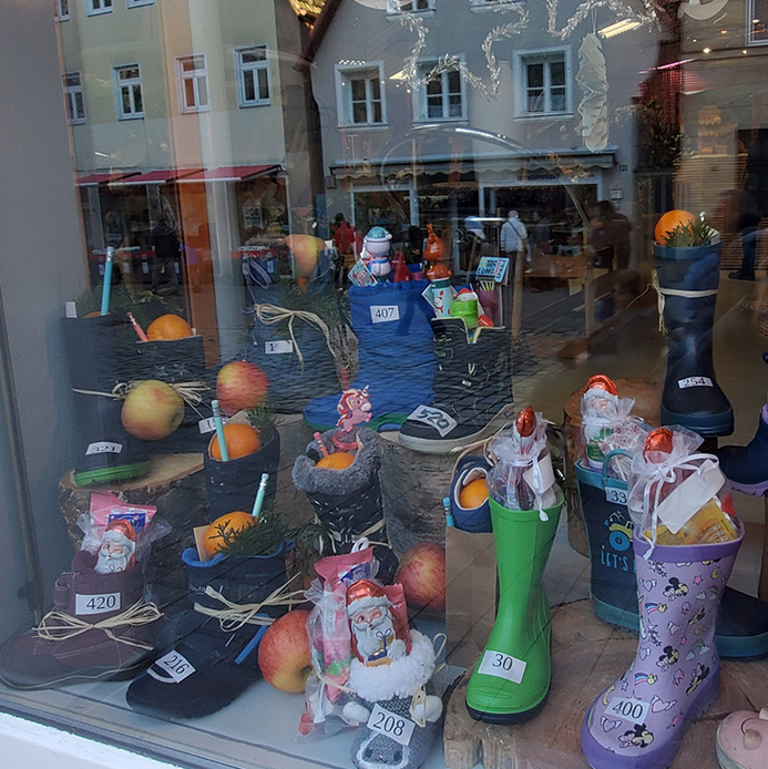 Im Schaufenster sind verschiedene bunte Weihnachtsstiefel aus Gummi und Stoff aufgestellt, die mit kleinen Geschenken, Süßigkeiten und Orangen gefüllt sind. Die Stiefel haben unterschiedliche Farben und Muster, darunter grün, lila mit Sternen, schwarz und rosa. Einige Stiefel tragen kleine Zettel mit Nummern. Im Hintergrund spiegeln sich Häuser und Passanten auf der Straße.