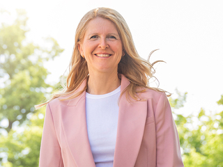 Blonde Frau im rosafarbenen Blazer mit weißem T-Shirt, im Hintergrund Bäume oder Sträucher