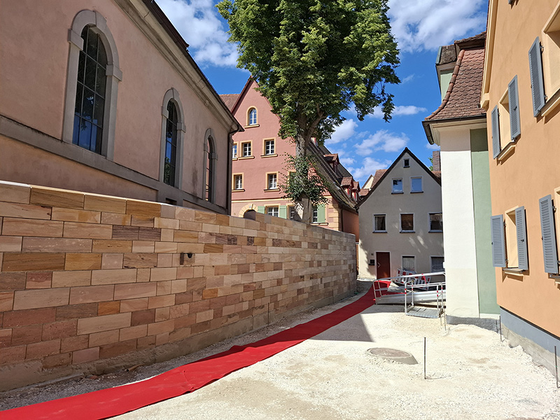 Eine neu errichtete Sandsteinmauer verläuft entlang einer Gasse zwischen Gebäuden. Der Boden ist noch nicht fertiggestellt, ein provisorischer roter Teppich markiert den Gehweg. Im Hintergrund sind Absperrgitter und Wohnhäuser zu sehen.