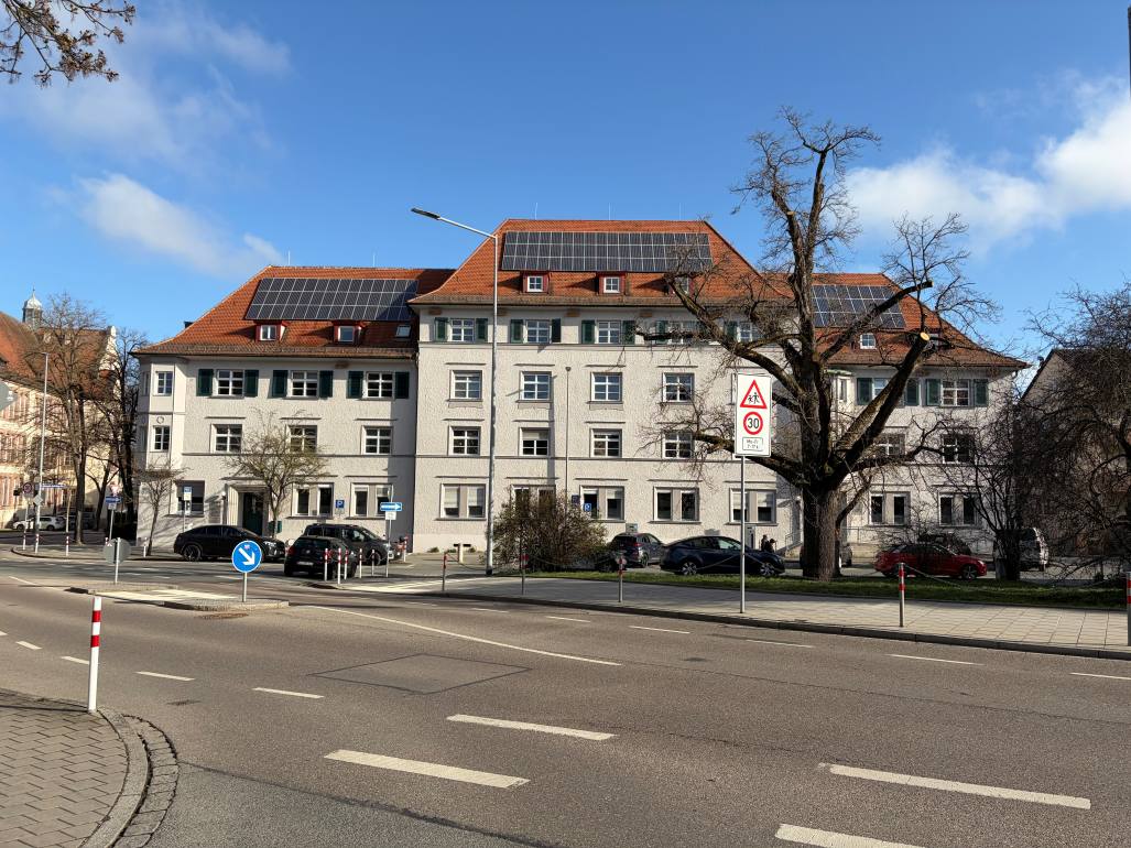 Eine breite Straße mit Fahrbahnmarkierungen und kleiner Verkehrsinsel führt an einem großen, hellen Gebäude mit rotem Ziegeldach und Solarpaneelen vorbei. Vor dem Gebäude stehen parkende Autos, daneben ein großer, unbelaubter Baum und ein Verkehrsschild mit Tempo 30 und Kinderwarnung. Der Himmel ist blau mit vereinzelten Wolken, die Umgebung wirkt ruhig und ordentlich.