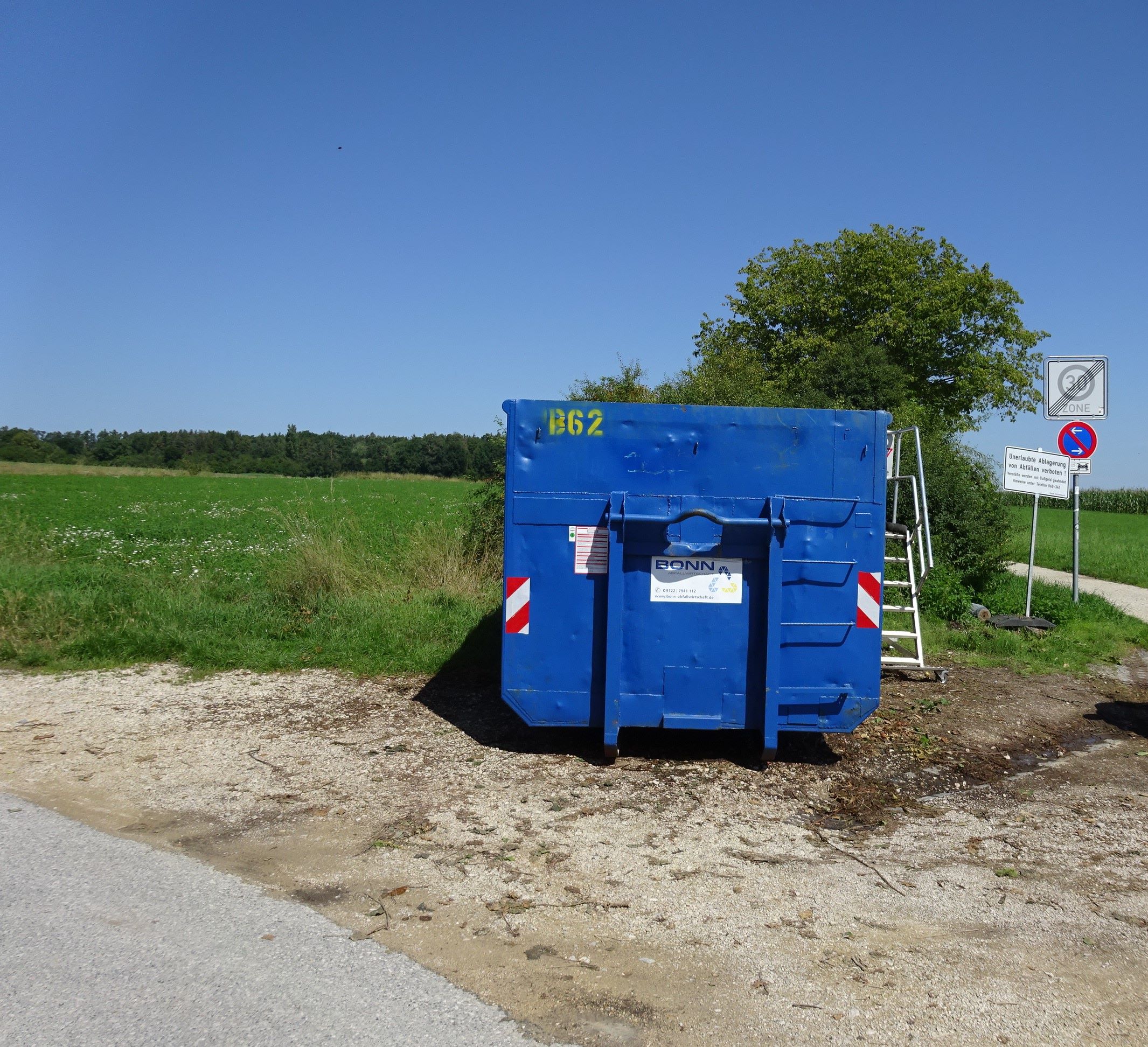 Ein blauer Abfallcontainer steht vor einer Wiese. Im Vordergrund sieht man eine sandige Straße und im Hintergrund sind eine Wiese, Bäume und Verkehrsschilder sowie ein blauer Himmel zu sehen. 