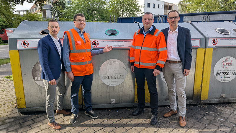 Vier Männer stehen nebeneinander vor einer Reihe von Glascontainern im Freien. Zwei der Männer tragen orangefarbene Warnwesten über Hemd und Hose, die anderen beiden sind in Anzügen gekleidet. Die Container sind mit runden Einwurföffnungen und klar beschrifteten Bereichen für Braunglas, Grünglas und Weißglas versehen. Auf jedem Container befindet sich ein deutlich sichtbarer Hinweis mit rotem Symbol, das das Einwerfen von Gegenständen außerhalb der Öffnungen untersagt. Im Hintergrund sind Bäume und ein Wohngebäude zu sehen.