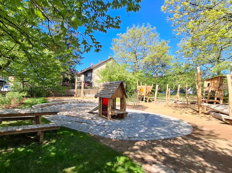 Blick auf einen Spielplatz mit einer Sandfläche und einem Spielhaus in der Mitte. Im Hintergrund sieht man ein Wohnhaus, vorne links eine Sitzgruppe aus Holz