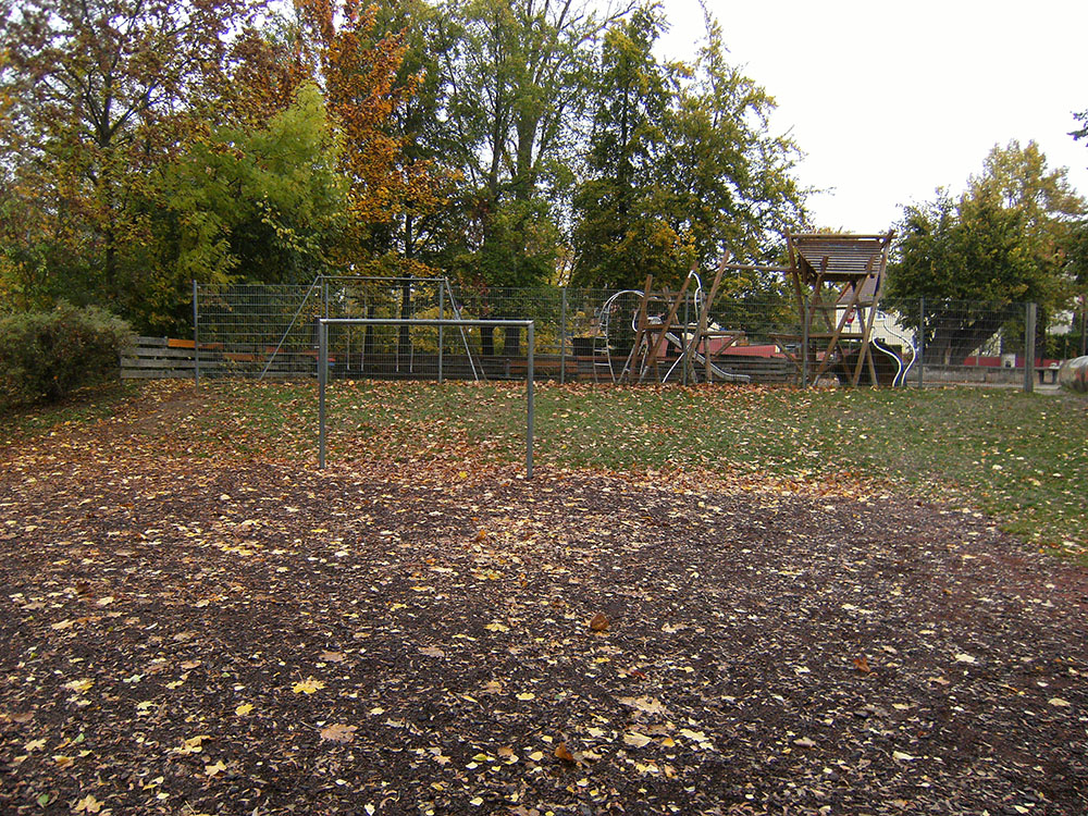 m Vordergrund liegt eine Fläche mit braunem Mulch, die von herabgefallenen gelben und braunen Blättern bedeckt ist. Dahinter steht ein Metallgestell, das wie ein Torrahmen aussieht. Im Hintergrund sind verschiedene Spielgeräte aus Holz und Metall zu sehen, darunter eine Kletterstruktur und eine Schaukel. Der Spielplatz ist von einem Zaun umgeben und von zahlreichen Bäumen eingerahmt, deren Laub herbstlich gefärbt ist.
