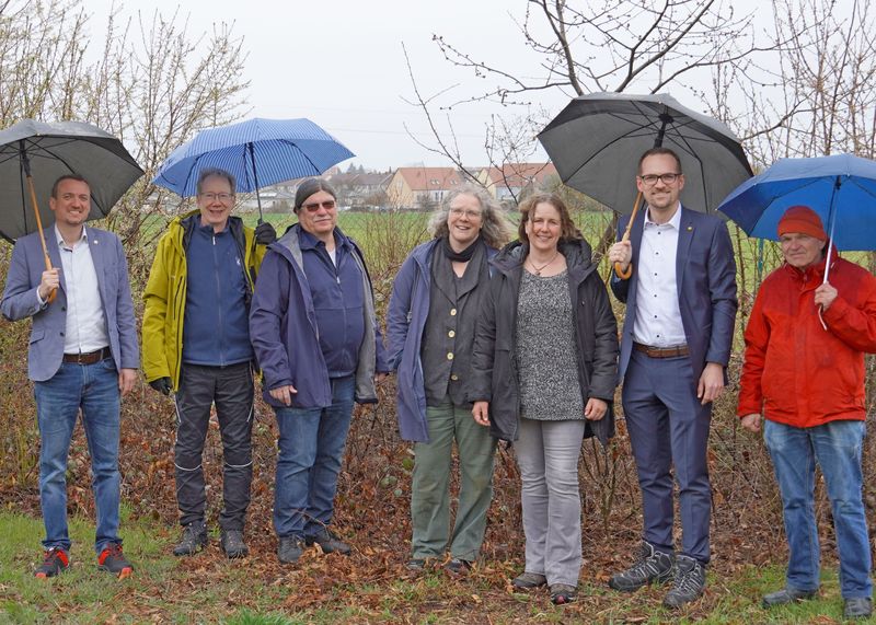Sieben Personen stehen auf einer Wiese mit Regenschirm. Es sind fünf Männer und zwei Frauen. Diese haben den Standort besichtigt.